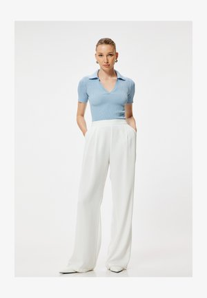 Mujer de pie con las manos en los bolsillos, vistiendo un top de punto azul claro de manga corta y pantalones anchos blancos de cintura alta sobre un fondo liso.