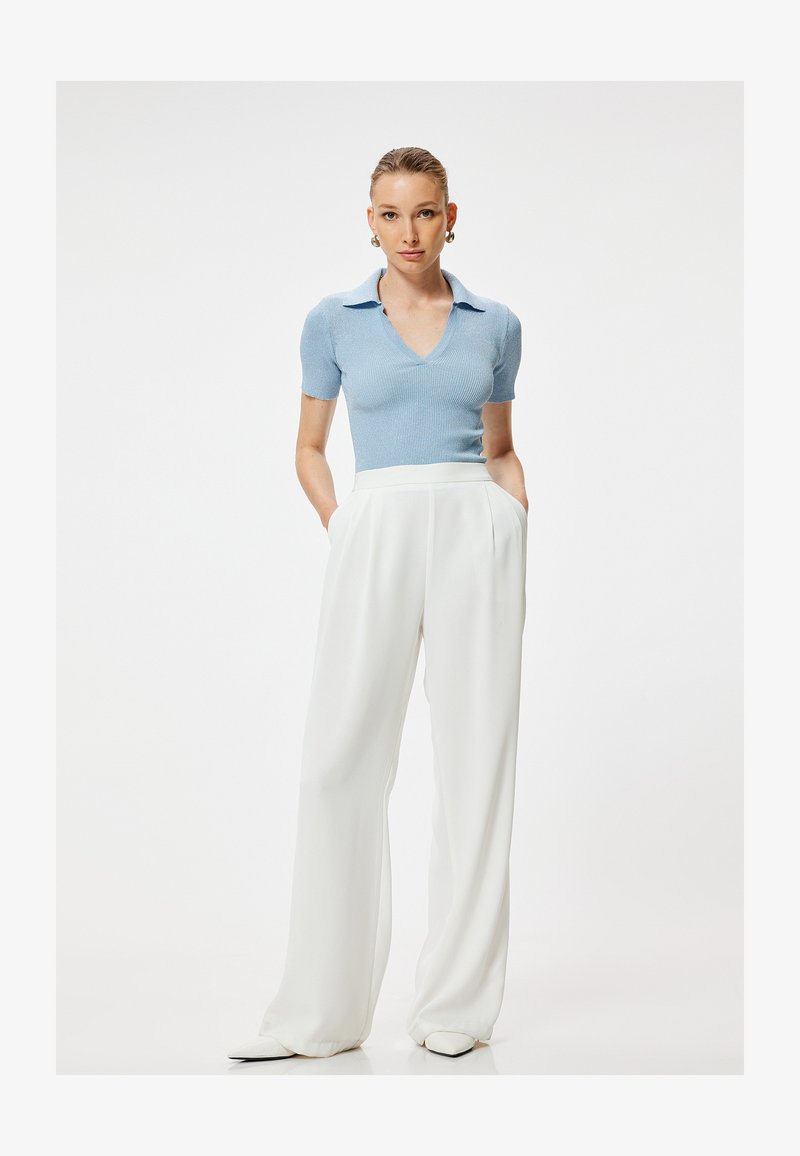 Kobieta stoi z rękami w kieszeniach, ubrana w jasnoniebieski krótki sweter z dzianiny i białe szerokie spodnie z wysokim stanem na jednolitym tle.