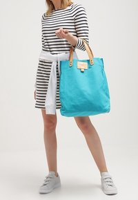 Stor turkos tygväska med beigea läderhandtag och en etikett. Bärs med en svartvit randig klänning och grå sneakers.