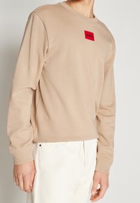 Felpa beige a maniche lunghe realizzata in tessuto morbido. Presenta una toppa rossa a forma rettangolare sul petto e polsini a coste.