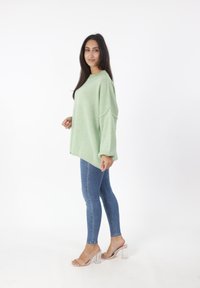Hellgrüner, übergroßer Pullover mit weicher Textur, kombiniert mit blauen Skinny-Jeans und klaren, absatzlosen Sandalen, vor einem schlichten weißen Hintergrund.
