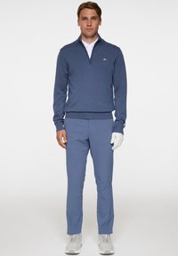 Miestens golf rūbai, kuriuose yra tamsiai mėlyna užtrauktuku užsegama megztinė ir atitinkantys kelnes, papildyti balta marškinėliais su apykakle ir pilkais sportiniais bateliais.