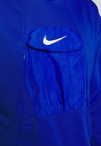 Sweat-shirt bleu avec une poche zippée contrastante de couleur bleue. Présente un logo Nike blanc sur la poche. Le tissu semble texturé.