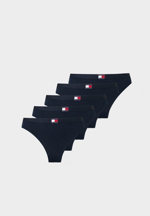 Γυναίκα που φοράει λευκό bralette Tommy Hilfiger και σκούρο μπλε εσώρουχα, στέκεται με το ένα χέρι στη μέση της μπροστά από μονόχρωμο φόντο.