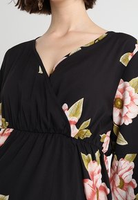 Černé šaty s zavinovacím designem, s květinovými vzory v růžové a zelené. Mají vytažený pas a dlouhé rukávy.