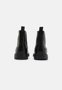 Bottes cheville en cuir noir à texture lisse, avec des détails décoratifs perforés et des semelles en caoutchouc. Équipées de languettes pour un enfilage facile.