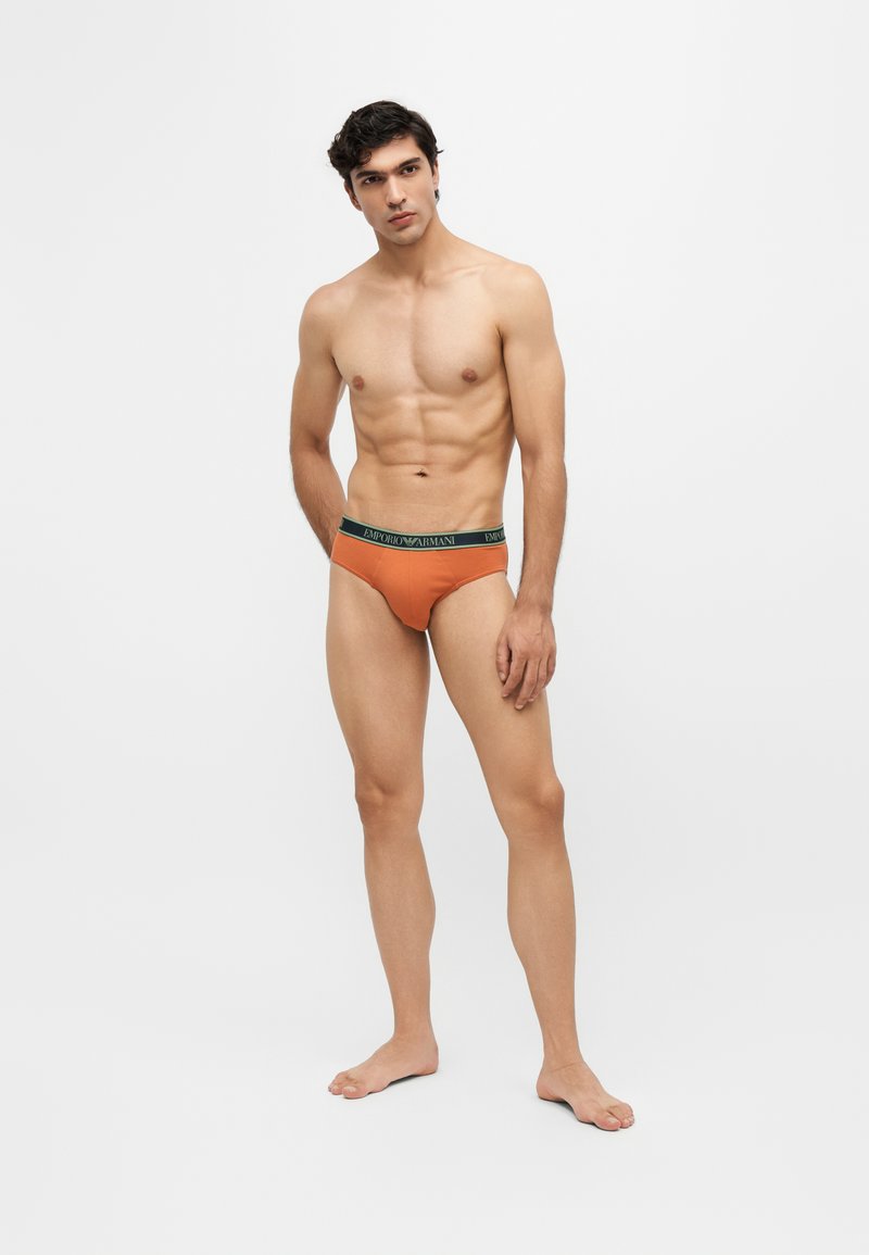 Jeune homme aux cheveux courts et foncés, debout pieds nus portant un slip orange Emporio Armani sur un fond blanc uni.