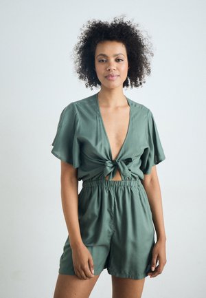 Grønn kortermet jumpsuit med v-hals og knytedetalj foran. Stoffet ser mykt og glatt ut, med elastisk midje for komfort.
