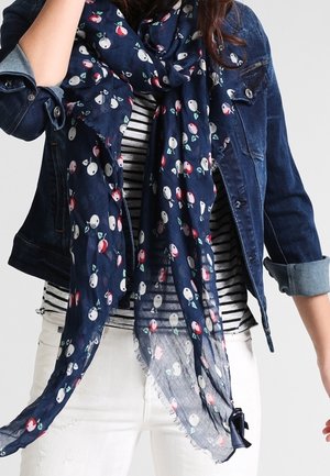 Femme portant une écharpe fleurie bleu foncé sur une chemise rayée noir et blanc, une veste en jean retroussée et un pantalon blanc.