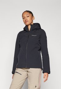 Schwarze wasserdichte Jacke mit Kapuze, Frontreißverschluss und verstellbaren Bündchen; mit Logo auf der Brust und kombiniert mit beigen Hosen.