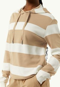 Sudadera a rayas beige y blanca con cordón, mangas largas, textura suave y un ajuste relajado. Diseño notable de rayas superpuestas.