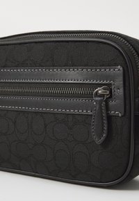 Schwarze Stofftasche mit strukturiertem Muster, ausgestattet mit einer vorderen Reißverschlusstasche und Lederakzenten. Glatter Reißverschluss mit einem dekorativen Ziehband.