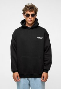 Zwarte hoodie met een grote voorzak en een capuchon met trekkoord, met witte tekst op de borst. Draagt het in combinatie met lichtblauwe spijkerbroek en een zonnebril.