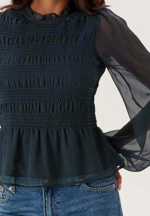 Blusa negra semitransparente de manga larga con cuerpo fruncido y bajo con volantes, combinada con jeans vaqueros azules.