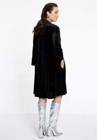 Longue robe en velours noir avec des manches trois-quarts et des poches latérales, associée à des bottes argentées métalliques jusqu'au genou avec un talon carré.