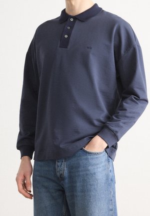 Granatowa koszulka polo z długim rękawem z kontrastującym kołnierzykiem, plisą na trzy guziki i subtelnym logo. Noszona z jasnoniebieskimi dżinsami.