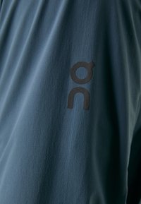 Korte mouwen sportshirt van donkerblauwe stof; heeft een gladde textuur en een prominent zwart logo op de linkermouw.