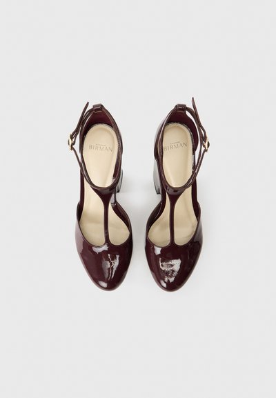 Bordowe lakierowane szpilki z wycięciami, pasek z detalami w złotym kolorze i zaokrąglonym noskiem. Gładka tekstura z obcasem typu słupek.