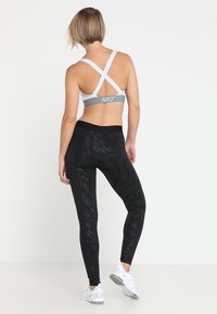 Sostenedor deportivo gris y blanco con tirantes cruzados, combinado con leggings negros que presentan un patrón sutil y zapatillas deportivas blancas.