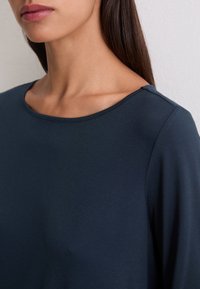 Donna con lunghi capelli scuri che indossa una maglietta a maniche lunghe blu scuro con colletto tondo, mostrata dal collo alle spalle.