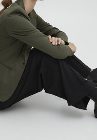 Zelený olivový střižený blejzr s hladkým textilním povrchem, doplněný širokými černými kalhotami. Jednoduchý design, žádné viditelné vzory ani kovové detaily.