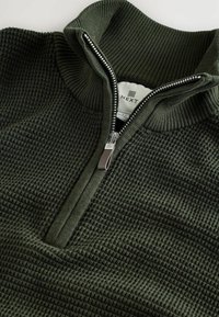 Suéter de punto texturizado en verde oscuro con media cremallera y cuello alzado acanalado, que muestra una etiqueta blanca de ropa en el interior del cuello.