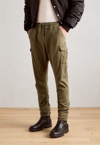 Olijfgroene cargo pants met een elastische tailleband, koord en zijzakken, gecombineerd met donkerbruine enkellaarzen op een houten vloer.