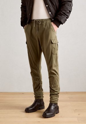 Olive green cargo pants with an elastic waistband, drawstring, and side pockets, paired with dark brown ankle boots on a wooden floor.