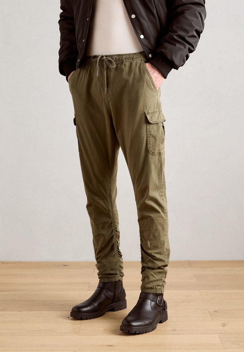 Olijfgroene cargo pants met een elastische tailleband, koord en zijzakken, gecombineerd met donkerbruine enkellaarzen op een houten vloer.