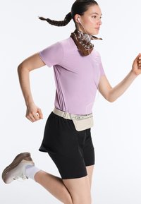 Ung person, der jogger iført en lilla skjorte, sorte shorts, beige bælte taske, brun halstørklæde, hvide øretelefoner og sneakers.