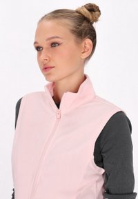 Gilet in pile rosa chiaro con colletto alto e chiusura con zip, indossato sopra una maglietta a maniche lunghe grigia scura. Texture liscia, design minimale.