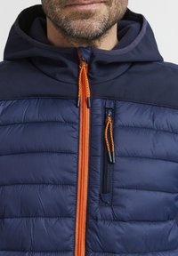 Marineblå pufferjakke med oransje glidelåser og snor, med en glidelåslomme på brystet og en myk hette. Strukturert overflate og isolert design.
