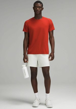 Rood atletisch T-shirt, gestructureerde stof, korte mouwen, gecombineerd met witte shorts. Houdt een witte waterfles vast met het "lululemon"-logo.
