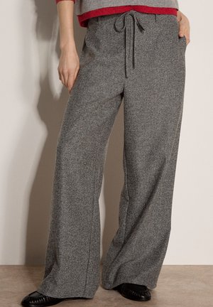 Pantalon gris à jambes larges avec taille à cordon, porté avec des chaussures noires et un pull gris et rouge visible à la taille.