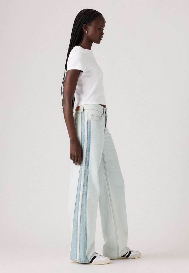 Weite, hellblaue Jeans mit rohem Saum und seitlichen Streifen. Kombiniert mit einem weißen Kurzarmhemd und weißen Sneakern mit schwarzen Streifen.
