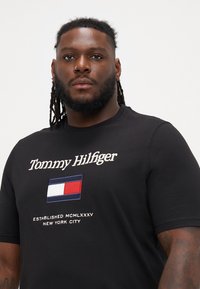 Musta puuvillane T-särk, millel on "Tommy Hilfiger" brodering, värviline lipu logo ja tekst "Asutatud MCMLXXXV New York City."