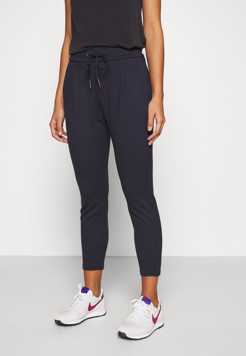 Personne portant un pantalon noir à cordon et des baskets blanches avec un swoosh rouge, debout devant un fond clair uni.