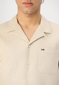 Beige kortermet skjorte laget av teksturert stoff. Har en notched krage, knappes front, og en venstre brystlomme med et stripete logo.