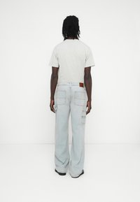 Világosszürke póló, világoskék, széles szárú farmernadrággal párosítva, amely több zsebbel és barna bőrpánttal rendelkezik a derékrészen.