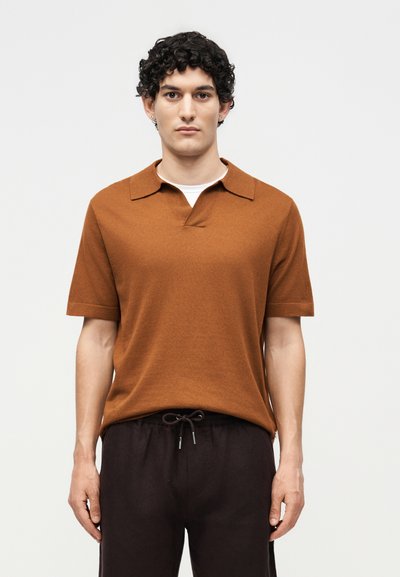 Homme aux cheveux bouclés portant une chemise marron à manches courtes avec col par-dessus un sous-vêtement blanc et un pantalon noir à cordon, debout devant un fond uni.