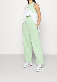 Un haut sans manches vert clair associé à un pantalon de survêtement léger vert clair. Un sweat-shirt blanc drapé sur les épaules, complété par des baskets blanches et grises.