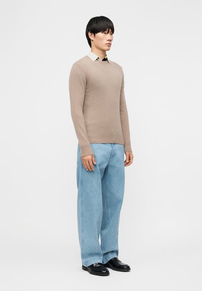 Νεαρός άνδρας που στέκεται, φοράει μπεζ πουλόβερ πάνω από λευκό πουκάμισο με γιακά, φαρδιά ανοιχτό μπλε τζιν και μαύρα παπούτσια, μπροστά σε απλό λευκό φόντο.