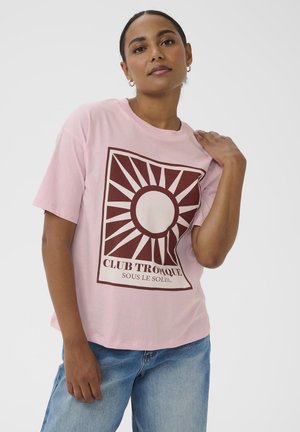 Femme portant un t-shirt rose clair avec un graphique de soleil rouge foncé et le texte "CLUB TROPIQUE SOUS LE SOLEIL", assorti à un jean bleu.