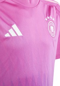 Różowy strój sportowy wykonany z gładkiego materiału, z wysokim kołnierzem, białym logo Adidas oraz okrągłym niemieckim emblematem narodowym z pięcioma gwiazdami.