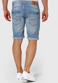 Pantaloni scurți din denim de culoare albastru deschis, cu tivuri rulotate, prevăzuți cu buzunare în spate cu clapete oblice și o etichetă cu logo pe talie. Adidași albi.