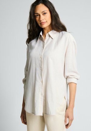Femme aux longs cheveux foncés portant une chemise ample crème claire boutonnée avec les manches retroussées et un pantalon clair, debout devant un fond uni.