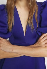 Henkilö, jolla on pitkät hiukset, pukeutuneena syvään V-pääntiehen lilaan mekkoon, jossa on puffimaiset hihat, kädet rinnan päällä.