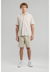 Camicia a maniche corte, a righe, beige, con taschino sul petto, abbinata a pantaloni corti color khaki e sneakers bianche. Design semplice, vestibilità rilassata.