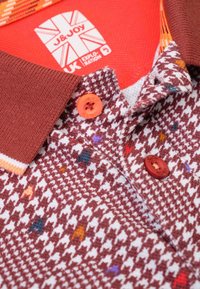 Geruite polo shirt in bruin en wit, met oranje knopen en kleurrijke gespikkelde accenten. Etiket toont merkl logo en informatie.