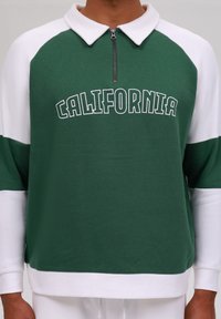 Zelený a biely sveter s golierom na zips, s nápisom "CALIFORNIA" v tučnom písmene, vyrobený z mäkkej látky s textúrovaným povrchom.